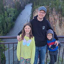 Michael Bicknell standing in front of a river on a bridge with two children smiling.