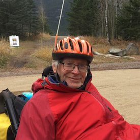 Bud is sitting on a wheel chair on a road, and he is wearing a red coat with an orange bicycle hat.