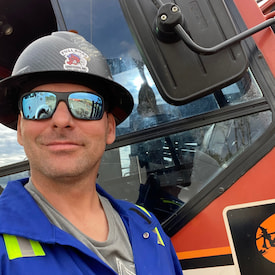 Jeremy is standing beside a large truck with a safty hat and a sun glasses.